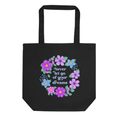 Motivational tote bag with quote: Never let go of your dreams. Text with blue and violet floral wreath with green foliage on black bag.